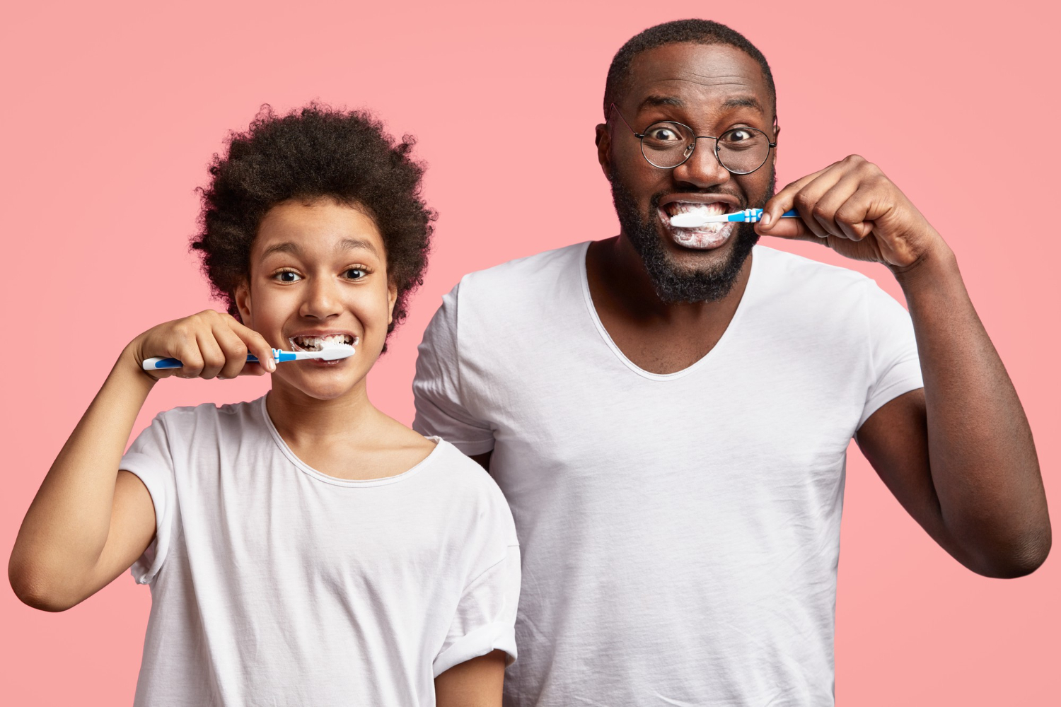 Fotografia colorida de homem e criança escovando os dentes. Ambos usam camisetas brancas e olhar animado.