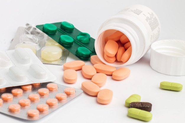 Fotografia colorida de pote de remédios derrubado, com diversas pílulas espalhadas. Além dessas, várias cartelas de remédios estão ao redor.