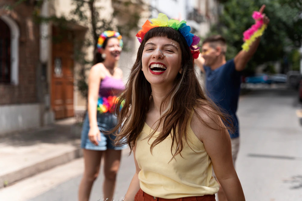 Mulher branca se diverte no carnaval enquanto usa acessórios coloridos. Atrás dela, outros foliões.
